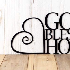 Close up of heart on our God Bless our Home metal sign, in matte black powder coat. Placed against a white wood wall.