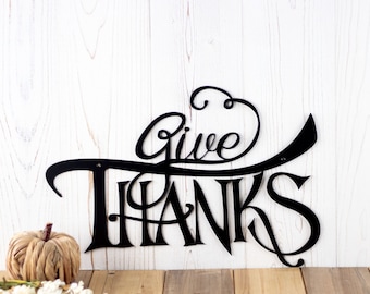 Könnte beinhalten: Ein schwarzes Metall-Wandschild mit der Aufschrift Give Thanks in einer stilisierten Schreibschrift. Das Schild hat eine glatte Oberfläche und ist vor einem weißen Holzmaserungshintergrund platziert, zusammen mit einem kleinen beigen Kürbis und weißen Blumen.