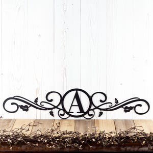 May include: Black metal monogram with a decorative scroll design and the letter "A" in a circle. The monogram is on a white wood background.