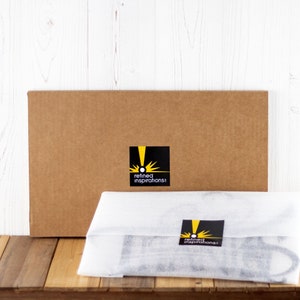 Cardboard folded box with Refined Inspirations sticker, displayed behind a custom metal sign, wrapped in white foam.