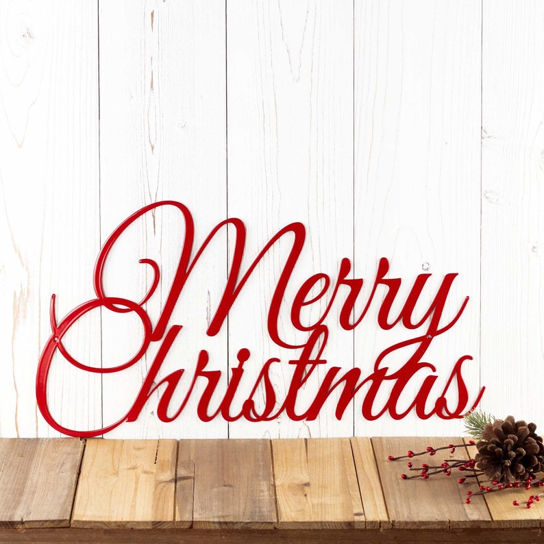 Merry Christmas script metal sign, in red gloss powder coat. Placed against a white wood wall.