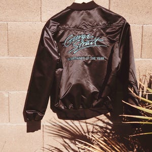 May include: Black satin bomber jacket with the text "George Strait" and "Entertainer of the Year" in teal script. The jacket is hanging on a wooden hanger against a brick wall.