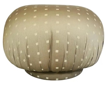 May include: A beige, upholstered ottoman with a rounded top and a circular base. The fabric is patterned with small, evenly spaced, square accents. The ottoman has a pleated design around the sides.