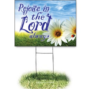 May include: A white yard sign with a blue sky and green grass background. The sign features a white daisy with a yellow center and a ladybug. The text on the sign reads "Rejoice in the Lord always".