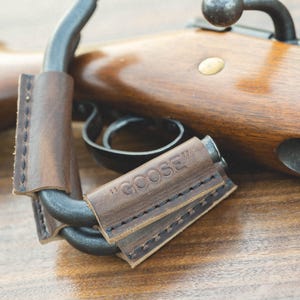 May include: A brown leather rifle sling with the word "GOOSE" embossed on it. The sling is attached to a rifle with a wooden stock.