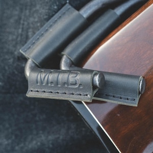 May include: Close-up of a black leather guitar strap with a metal buckle and the text "MTB" embossed on the strap.