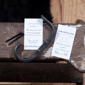 May include: Black metal hook with instructions for mounting and care. The instructions include the text "Mount directly into wood or wall stud (recommended)" and "Simply clean and protect as needed using a small amount of Wax or Oil".