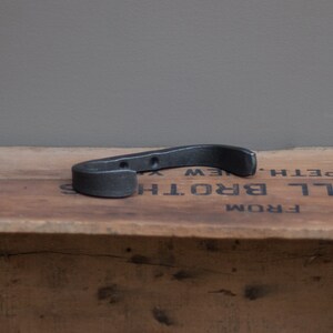 May include: A black metal hook with a curved shape. The hook is sitting on a wooden surface with the text "FROM LL BROTH'S NEW  BETH'" visible.