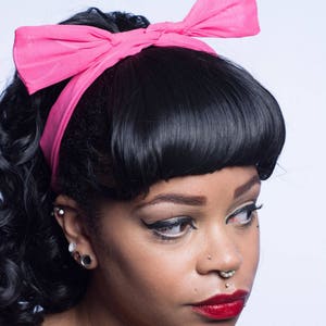 Pode incluir: Uma mulher com cabelo preto preso em um rabo de cavalo com uma faixa de cabeça rosa. Ela está usando maquiagem com delineador de olhos alado e batom vermelho. Ela tem várias piercings nas orelhas e no nariz.