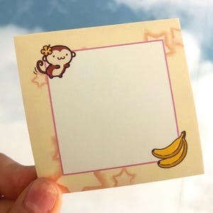 May include: A square, light yellow sticky note with a pink border. The note features a cartoon monkey with a flower and a pair of bananas. The background has a star pattern. The note is held by a hand.