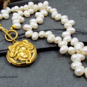 May include: A pearl necklace with a gold-colored pendant. The pendant features a detailed face design. The necklace is composed of numerous white pearls and has a gold clasp. The necklace is displayed on a dark gray surface.