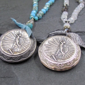 May include: Two silver pendant necklaces. Each pendant features a woman with outstretched arms, sun rays, and a decorative border. One necklace has turquoise beads, the other white. Both have ribbon accents.