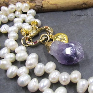 May include: A white pearl necklace with a gold clasp and a purple amethyst pendant. The necklace features a mix of round and irregular pearls. The clasp and pendant accents are gold-colored. The pendant is a raw, natural amethyst crystal.