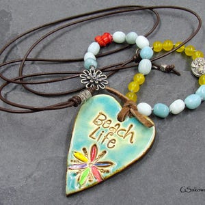 May include: A handcrafted necklace featuring a turquoise heart-shaped pendant with the words "Beach Life" and a colorful flower design. The necklace includes a brown leather cord and beaded accents in various colors.