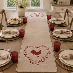 May include: A long, rectangular beige table runner made of woven fabric featuring a red printed design of a rooster centered within a heart-shaped wreath of leaves and branches, with a smaller matching rooster wreath motif at the top end.