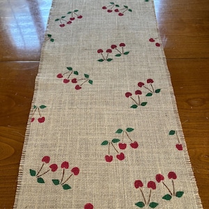 May include: A burlap table runner with a red cherry pattern. The cherries are printed on a natural burlap fabric with a frayed edge.