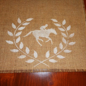 Pu&ograve; includere: Un runner da tavolo in juta con un design a stencil bianco di un cavallo e un cavaliere in una corona di alloro.