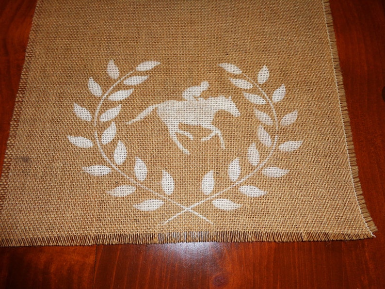 Pu&ograve; includere: Un runner da tavolo in juta con un design a stencil bianco di un cavallo e un cavaliere in una corona di alloro.