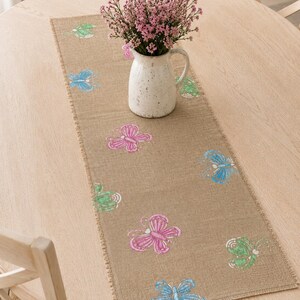 Puede incluir: Un camino de mesa rectangular de arpillera color canela con un patrón repetitivo de mariposas impresas en tonos rosa, azul y verde. El camino tiene bordes deshilachados y está coronado por una jarra de cerámica blanca con flores rosas.