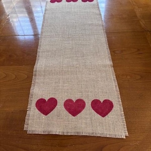Puede incluir: Camino de mesa rectangular de arpillera con borde deshilachado. El camino de mesa presenta tres corazones burdeos en cada extremo y tres corazones más pequeños en el centro, creando un acento decorativo.