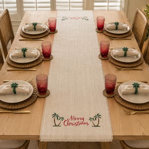 May include: A light beige linen table runner featuring embroidered green palm trees and the red text Merry Christmas at both ends, centered on a wooden dining table set with white plates, red textured glasses, and green palm tree napkin rings.