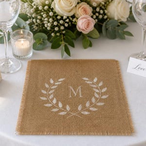 May include: A square burlap placemat with frayed edges featuring a white printed design of a laurel wreath surrounding the capital letter M centered on the fabric. The placemat is positioned on a white tablecloth with a floral arrangement nearby.