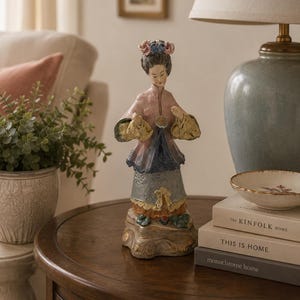 May include: A ceramic figurine of a person wearing a pink jacket with yellow sleeves, a dark blue vest, and a grey skirt. The figure has dark hair styled with pink and blue floral accents, standing on a decorative base with gold-toned detailing.