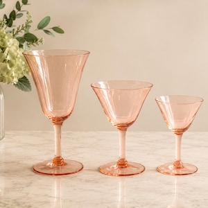 Pode incluir: Três taças de vidro rosa claro de tamanhos diferentes dispostas em fila. Cada taça apresenta um acabamento liso e translúcido, uma haste fina com um pequeno nó decorativo na base e um pé circular largo para estabilidade na superfície.