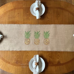 Könnte beinhalten: Ein Tischläufer aus Jute mit einem Ananas-Design. Der Läufer liegt auf einem Holztisch mit vier weißen Tellern und Holz-Serviettenringen.