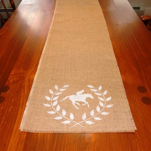 Pu&ograve; includere: Un runner da tavolo in juta marrone con un design a cavallo bianco e corona di alloro. Il runner &egrave; su un tavolo di legno.