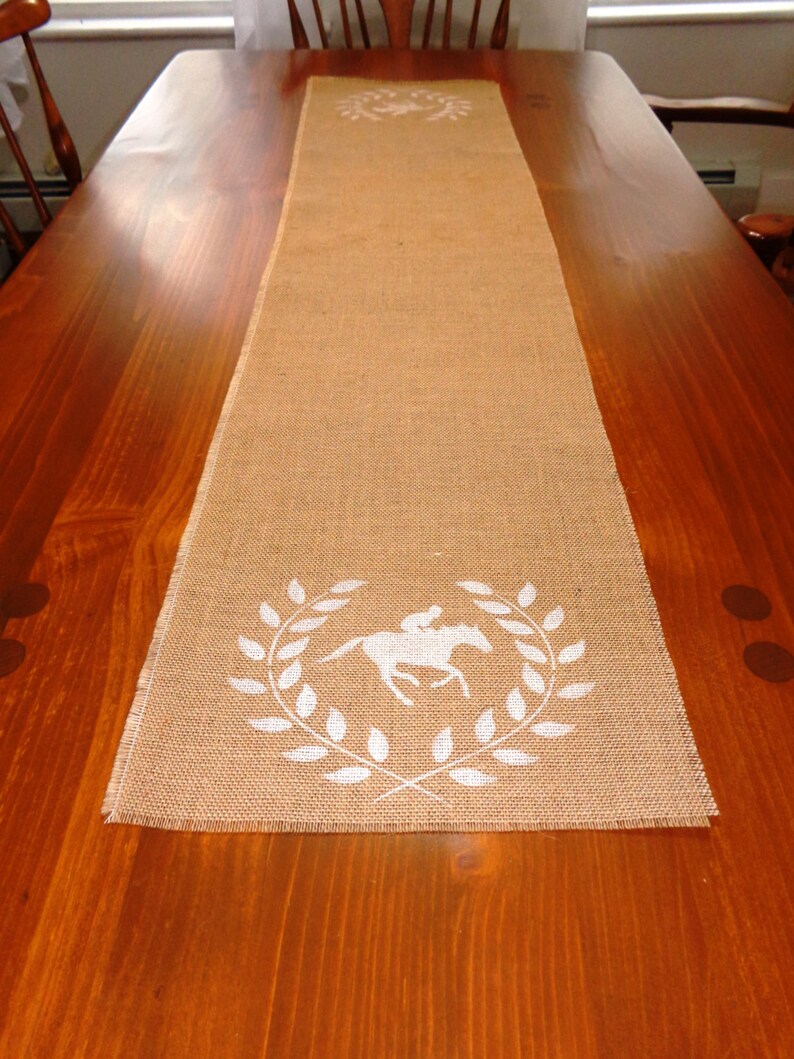 Pu&ograve; includere: Un runner da tavolo in juta marrone con un design a cavallo bianco e corona di alloro. Il runner &egrave; su un tavolo di legno.