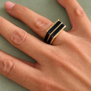 May include: A gold-toned ring featuring a wide, flat-top band with two parallel, rectangular black enamel inlays running horizontally across the center. The metal edges are polished, creating a distinct contrast with the smooth, dark inlay.