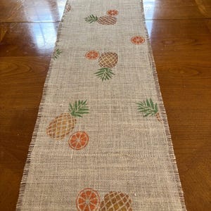 May include: A burlap table runner with a pineapple and orange design. The runner is beige with green leaves and orange fruit printed on it.