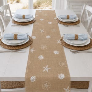 May include: A tan burlap table runner featuring a white printed pattern of starfish, seashells, and sand dollars. The runner has frayed edges and is centered on a white table with two place settings, each including a woven placemat and napkin.