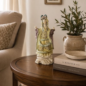 May include: A ceramic figurine of a person wearing a pale yellow robe with purple sleeves and intricate floral detailing. The figure has dark hair styled with pink flowers and stands on a sculpted base with flowing, wave-like patterns in cream.