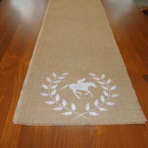 Pu&ograve; includere: Un runner da tavolo in juta marrone con un design bianco di un cavallo e un cavaliere in una corona di alloro.