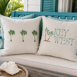 May include: Two square off-white throw pillows with green palm tree graphics. One pillow features three individual palm trees, while the other displays two palm trees next to the blue and green text that reads KEY WEST in a stacked format.