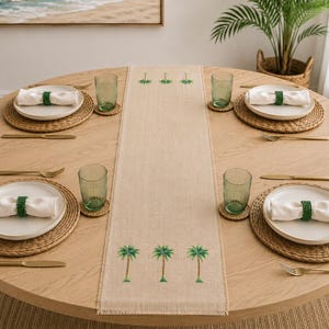 May include: A light beige linen table runner featuring embroidered green palm tree motifs, centered on a round wooden dining table. The setting includes four white plates on woven placemats, green ribbed glasses, and white napkins with green rings.