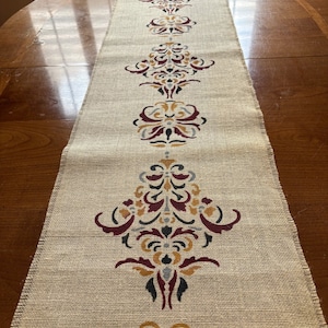 May include: A rectangular burlap table runner with a repeating pattern of ornate floral designs in burgundy, gold, gray, and black. The runner has a frayed edge and is displayed on a wooden table.