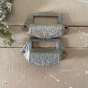 May include: Two antique drawer pulls with a decorative floral design. The pulls are made of metal and have a gray finish.