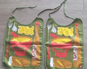 May include: Two green bibs with a food-themed design. Each bib features a red fondue pot, a cheese wedge, a bottle of wine, a baguette, a fork, and garlic. The bibs have green ties at the top.