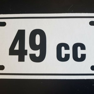 May include: White rectangular sign with black rounded corners and black text that reads "49 cc".