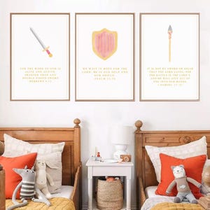 May include: Three framed watercolor prints with religious themes. The prints feature a sword, shield, and spear, each with a corresponding Bible verse in gold lettering. The artwork is displayed above two wooden beds with orange pillows and stuffed animals.
