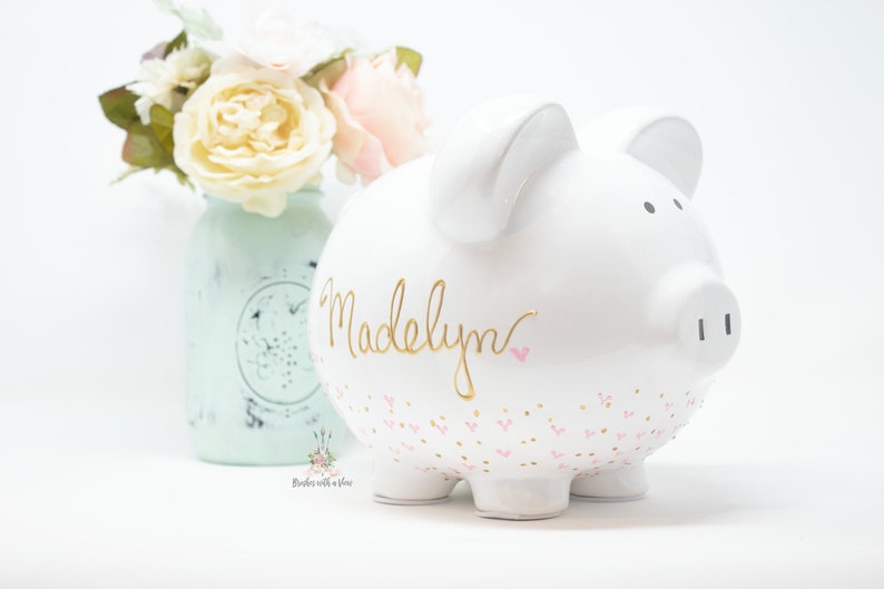 May include: White ceramic piggy bank with a gold inscription that reads "Madelyn" and pink and gold heart accents.