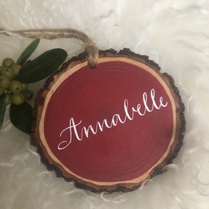 May include: A red wooden ornament with a natural wood bark edge. The ornament is painted red and has the name "Annabelle" written in white cursive lettering.