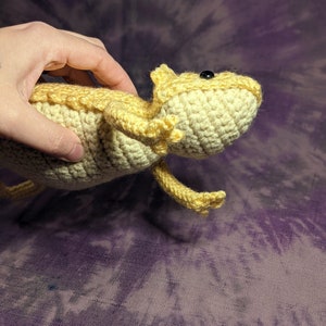 May include: A crocheted bearded dragon plush toy in a light yellow colour. The toy has black eyes and is posed with its head tilted up.
