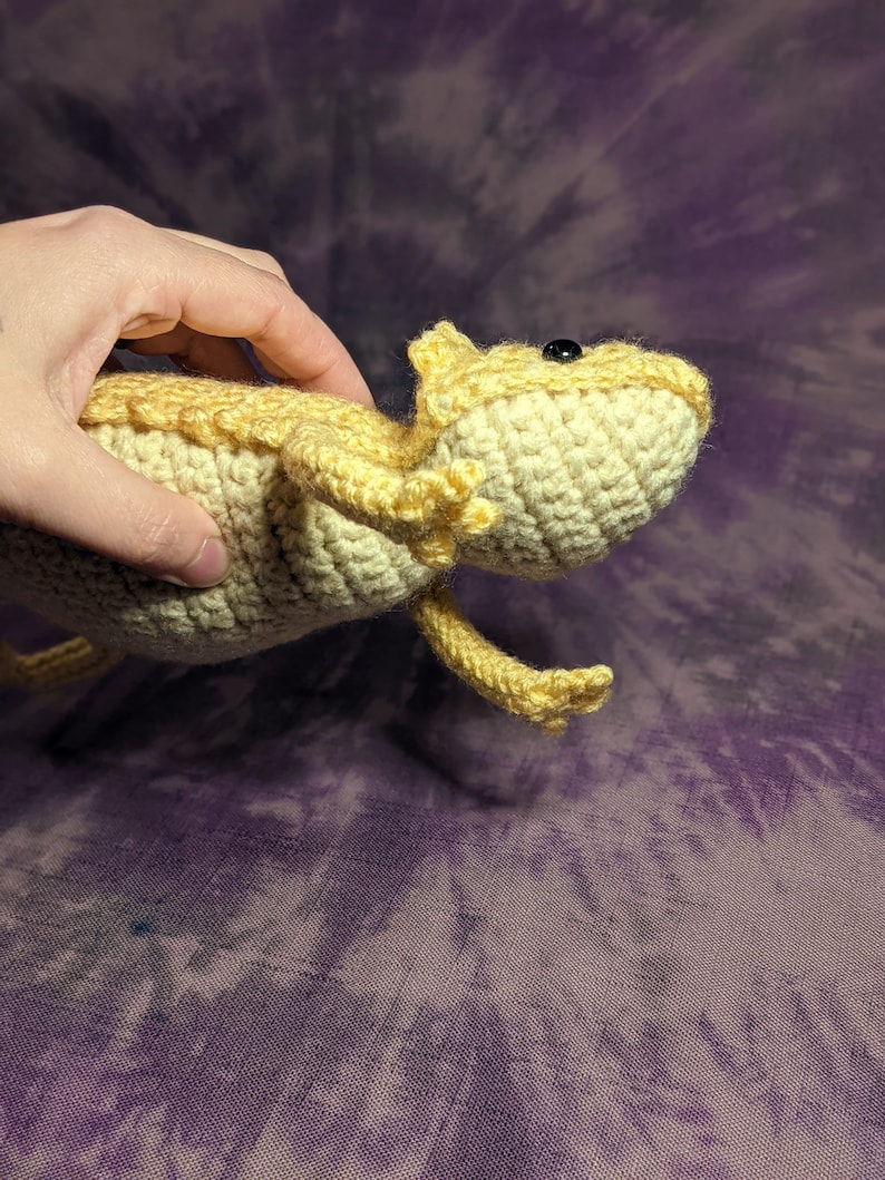 May include: A crocheted bearded dragon plush toy in a light yellow colour. The toy has black eyes and is posed with its head tilted up.