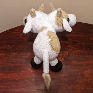 May include: A crocheted two-headed cow plush toy with white and brown fur, black eyes, and black hooves. The toy is sitting on a wooden table.