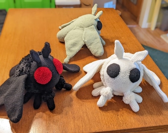 Op de afbeelding: Drie gehaakte motten knuffels in verschillende kleuren. Eén is zwart met rode ogen, één is wit met zwarte ogen en één is groen met zwarte ogen.