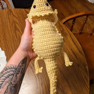 May include: A crocheted yellow bearded dragon plush toy with black eyes. The toy is being held by a hand with a tattoo.